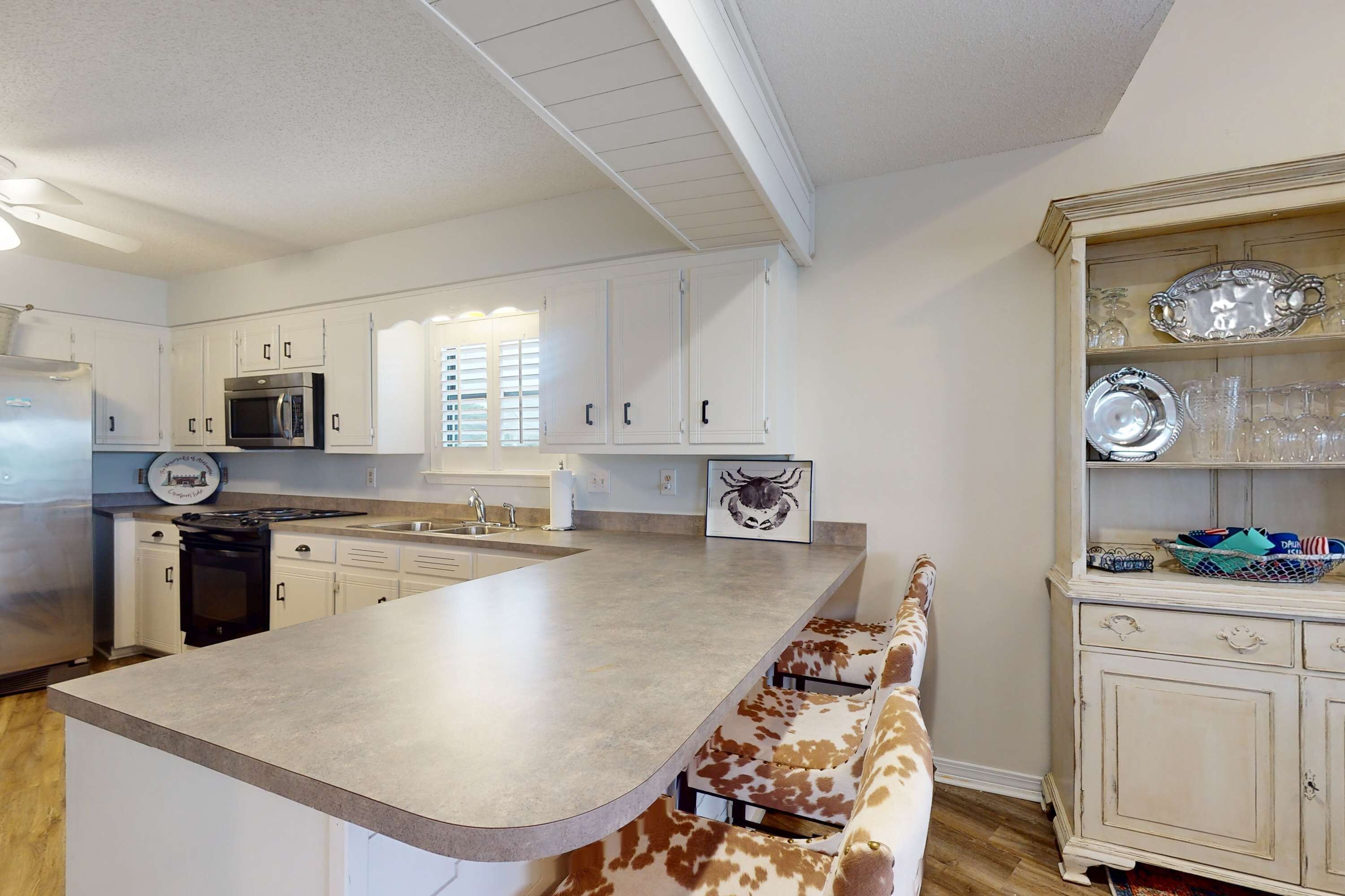 A kitchen with white cabinets, a stainless steel refrigerator, a microwave, and a large island with a cow-print bench.