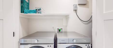 A laundry area with two Samsung washing machines, a shelf with toiletries, and a wall-mounted heater.