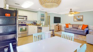Cocina con armarios blancos, microondas y cafetera. Hay una mesa de comedor con sillas y un sofá con cojines naranja.