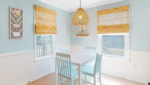Un comedor con una mesa y sillas blancas, una lámpara colgante y ventanas con persianas de bambú.