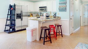 Cocina con armarios blancos, refrigerador de acero inoxidable, microondas y cafetera. Hay dos taburetes y una escalera.