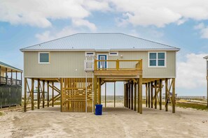 Ein zweistöckiges Strandhaus mit Balkon, Holzterrasse und Sandboden.