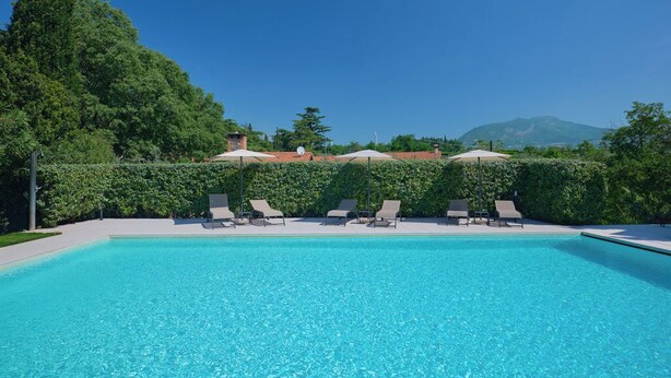 Ein klares, blaues Schwimmbecken mit Liegestühlen und Sonnenschirmen, umgeben von viel Grün und einem Berg im Hintergrund.