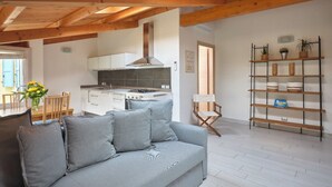 Ein modernes Wohnzimmer mit grauem Sofa, Holzbalken, einer Küche mit weißen Schränken und einem Bücherregal aus Holz.