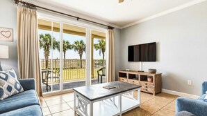 A living room with a sofa, a coffee table, a TV, and a sliding glass door leading to a balcony with palm trees. - Perfect beachfront condo with pool, internet, and washer and dryer (Dauphin Island)