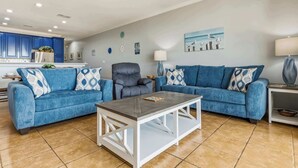 A living room with two blue sofas, a coffee table, a grey armchair, and a kitchen in the background. - Perfect beachfront condo with pool, internet, and washer and dryer (Dauphin Island)