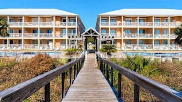 A hotel with a wooden boardwalk leading to a pool area, featuring multiple balconies and a clear sky. - Perfect beachfront condo with pool, internet, and washer and dryer (Dauphin Island)