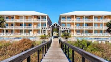 A hotel with a wooden boardwalk leading to a pool area, featuring multiple balconies and a clear sky.