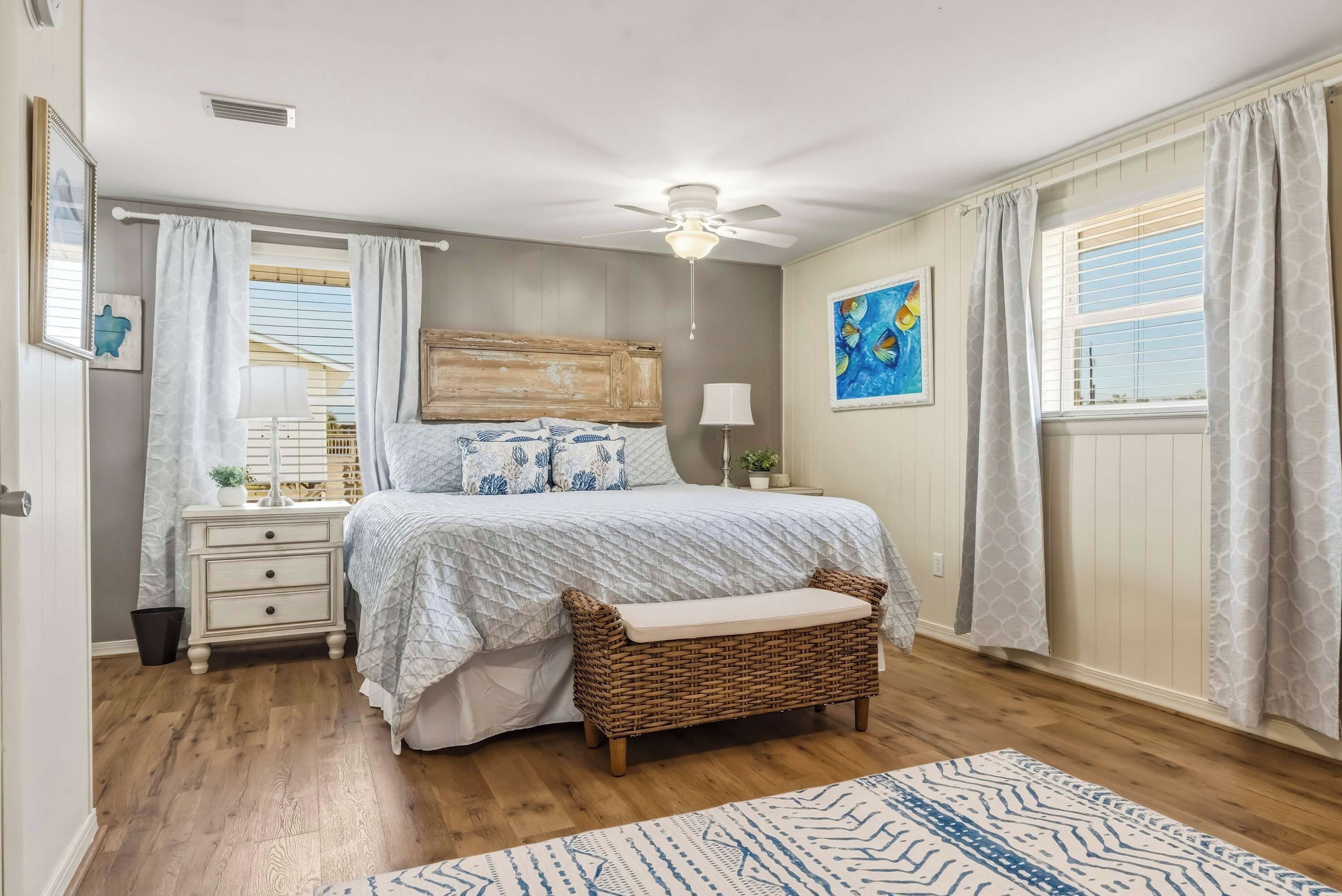 Une chambre à coucher comprenant un lit, des tables de chevet, un ventilateur de plafond, une fenêtre avec des rideaux, un tableau accroché au mur et un banc en osier.