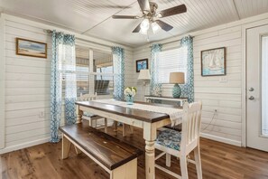 A dining area with a wooden table, white chairs, a ceiling fan, and patterned curtains. - Dog-friendly house on Dauphin Island, Near beach access, Patio, Grill, Washer & Dryer (Dauphin Island)