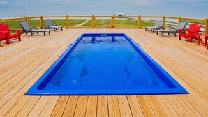 Um deck de madeira com piscina retangular, cadeiras de descanso e vista para a praia.