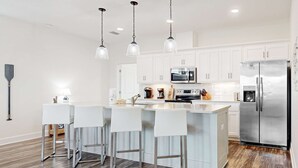 A modern kitchen with white cabinets, stainless steel appliances, and a central island with bar stools. - Coastal dog-friendly home with grill, balcony, & patio - short walk to the beach (Dauphin Island)