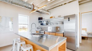 Una cocina moderna con electrodomésticos de acero inoxidable, taburetes de bar blancos y un ventanal grande.