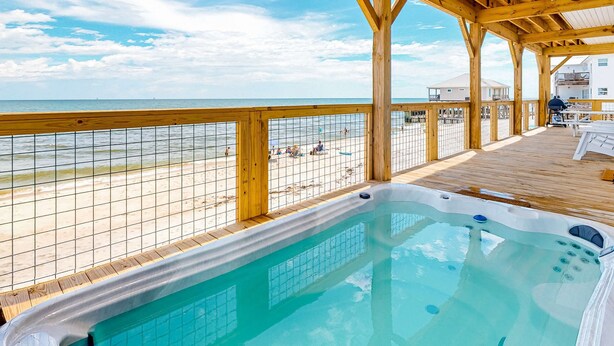 Una terraza de madera con piscina, vista a la playa y un área techada con mesas y sillas.