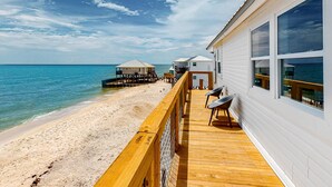 Una terraza de madera con barandilla da vista a una playa, un edificio con ventanas grandes y una estructura con techo de paja.