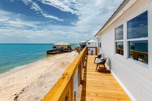 A wooden deck with a railing overlooking a beach, a building with large windows, and a structure with a thatched roof. - Gulf Front Paradise - Swim Spa, Dolphin Spotting, and Beach at Your Doorstep (Dauphin Island)