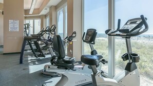 A gym with stationary bikes, treadmills, and an elliptical machine, with large windows offering a view of the outdoors. - Coastal Condo w Balcony & Shared Pools, Hot Tubs, Gym and Gulf views (Gulf Shores)
