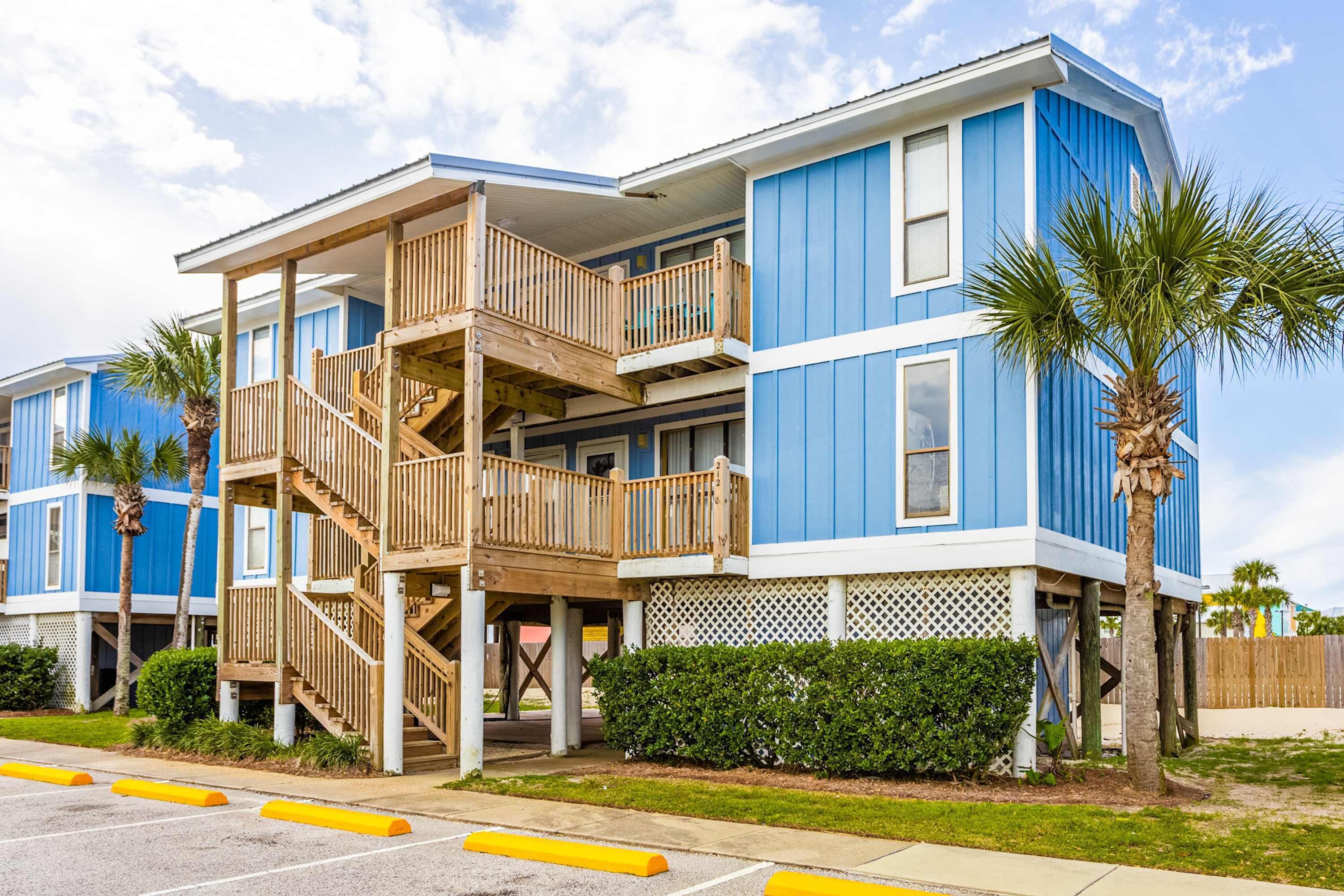 A multi-story building with a wooden balcony, a palm tree, and a fenced yard.