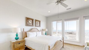 A bedroom with a large bed, two nightstands, a ceiling fan, and a view of the beach through large windows. - Beautiful gulf-front beach home with sweeping views from balcony, shared pool, WiFi (Gulf Shores)