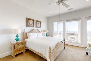 Ein Schlafzimmer mit einem großen Bett, zwei Nachttischen, einem Deckenventilator und Blick auf den Strand durch große Fenster.