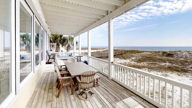 A covered outdoor deck with a table and chairs, overlooking a beach. - Beautiful gulf-front beach home with sweeping views from balcony, shared pool, WiFi (Gulf Shores)