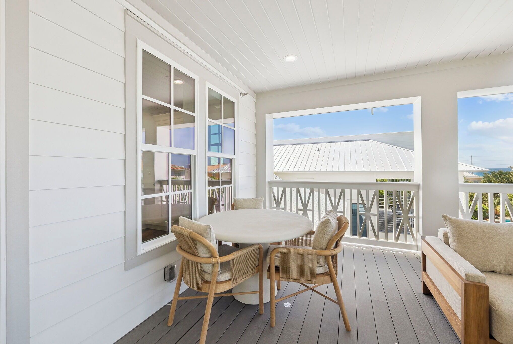 A covered patio with a round table and wicker chairs, a white railing, and a view of a building with a white roof.