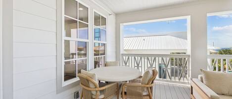 A covered patio with a round table and wicker chairs, a white railing, and a view of a building with a white roof.