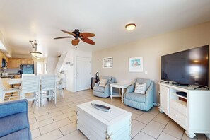 A living room with a TV, a ceiling fan, a dining area with chairs, and a kitchen in the background. - Gulf-front haven with free WiFi, central AC, & balcony - snowbirds OK (Gulf Shores)