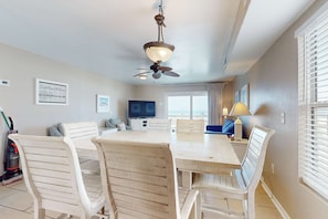 A dining area with a table and chairs, a ceiling fan, a television, and a window with blinds. - Gulf-front haven with free WiFi, central AC, & balcony - snowbirds OK (Gulf Shores)