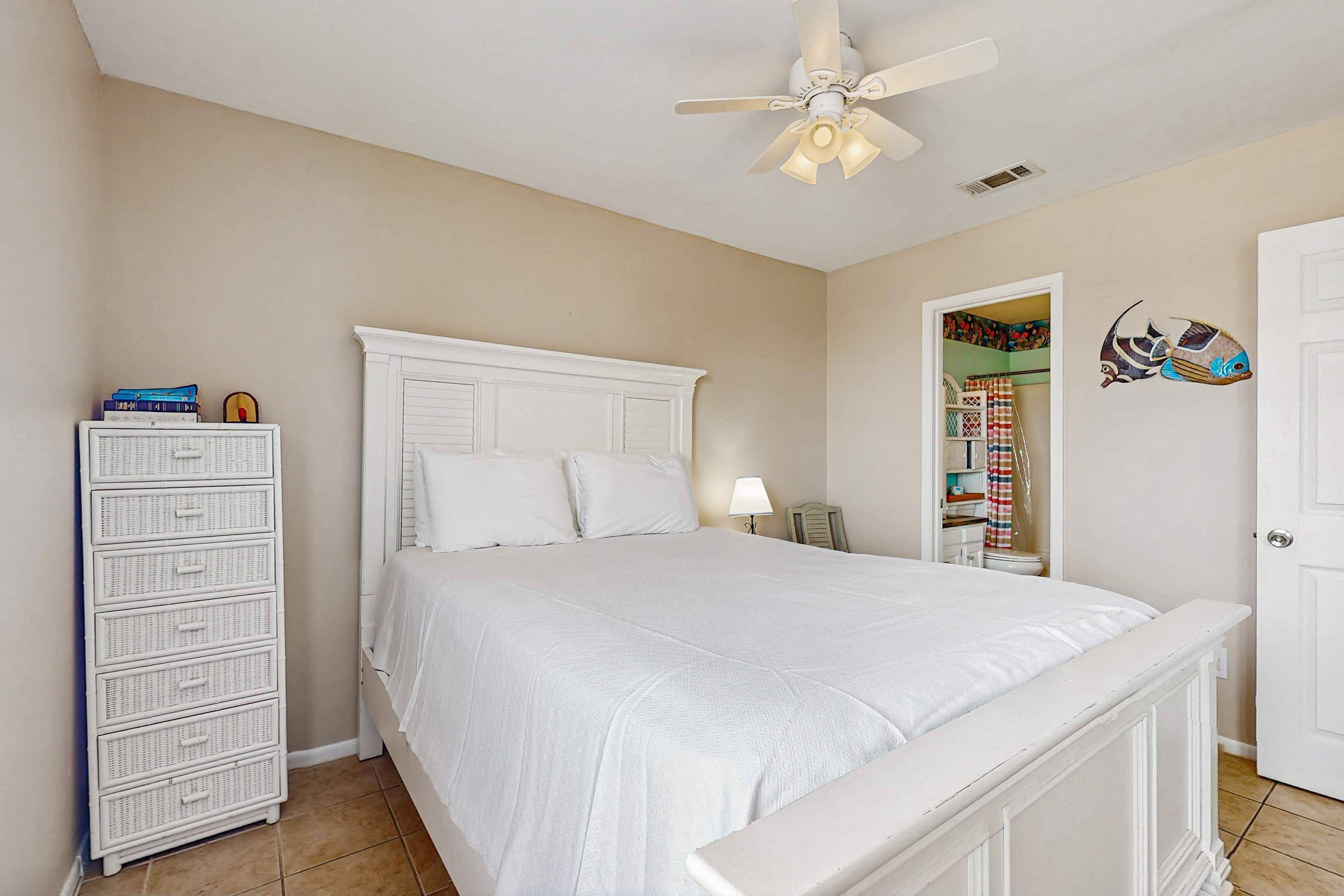 Une chambre avec un lit blanc, une commode blanche, un ventilateur de plafond et une porte décorée avec un motif de poisson.