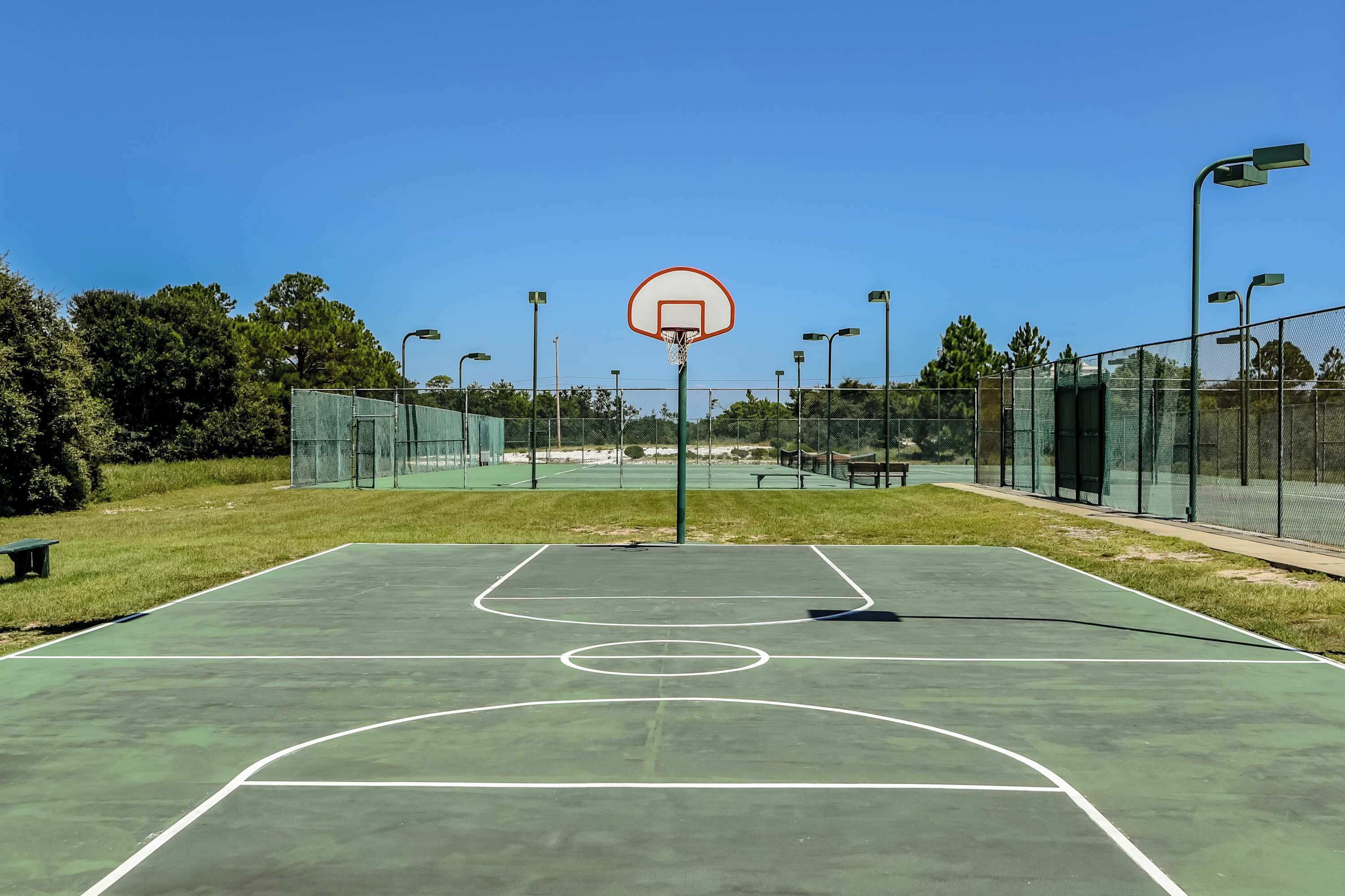 Ein Basketballplatz im Freien mit Netz, umgeben von einer Umzäunung, Bänken und Straßenlaternen.