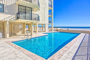 A swimming pool with a clear view of the ocean, a building with balconies, and a tiled deck area. - Bright, comfortable condo in beachfront complex with pool, central AC, & balcony (Gulf Shores)