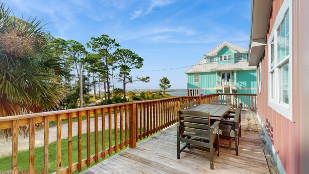 Une terrasse en bois avec une table et des chaises, d’où l’on peut admirer un bâtiment turquoise et une plage.