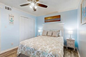 A bedroom with a bed, a ceiling fan, a nightstand, a wall-mounted painting, and a door. - Coastal condo just steps from the beach w shared pool, BBQ area, WiFi, AC, W&D (Gulf Shores)