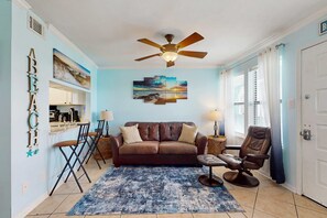 A living room with a brown leather sofa, a blue patterned rug, a ceiling fan, and a painting of a sunset. - Coastal condo just steps from the beach w shared pool, BBQ area, WiFi, AC, W&D (Gulf Shores)