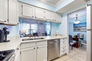 A kitchen with white cabinets, a dishwasher, and a dining area with a table and chairs. - Coastal condo just steps from the beach w shared pool, BBQ area, WiFi, AC, W&D (Gulf Shores)