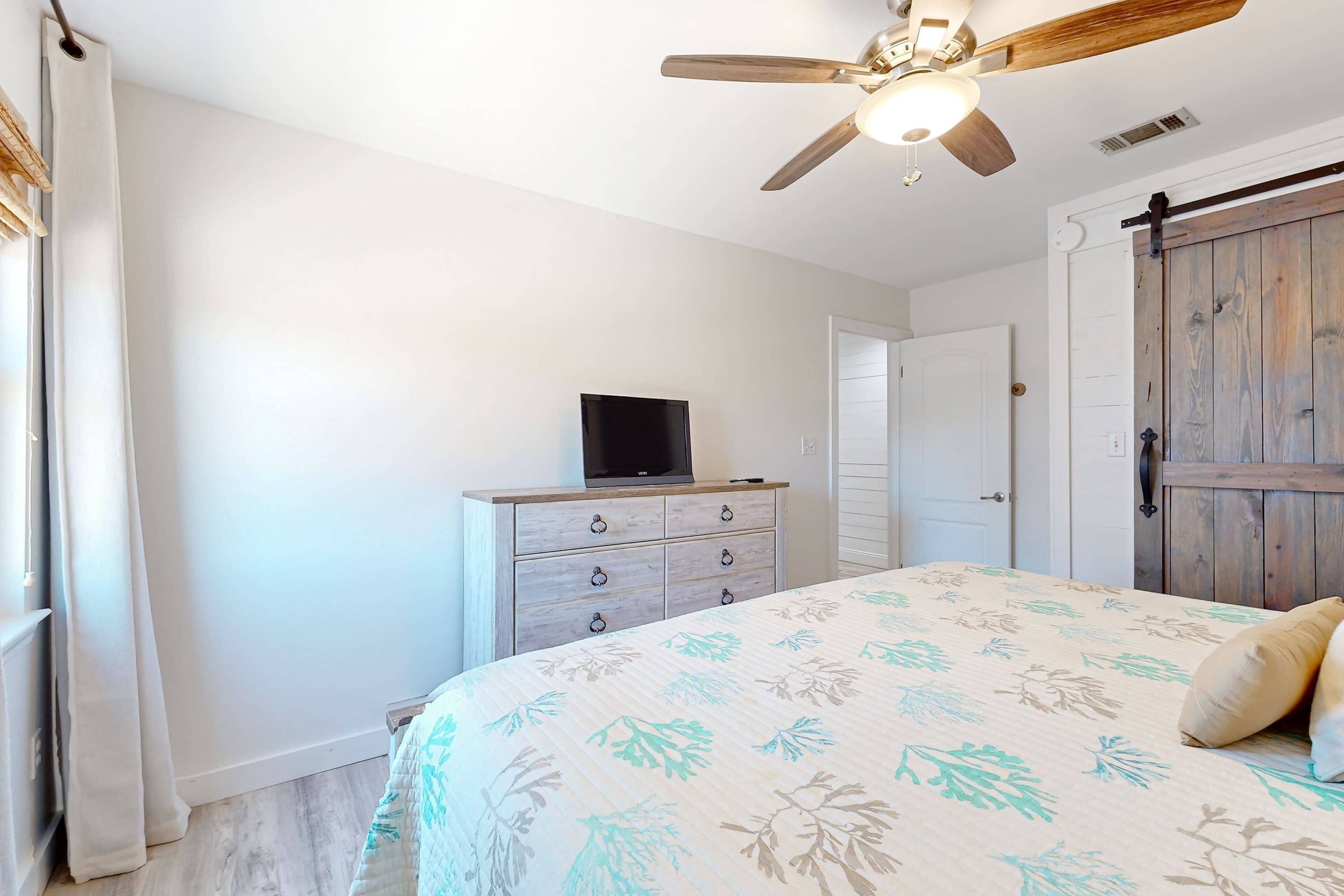 A bedroom with a bed, a ceiling fan, a television on a dresser, and a wooden door.