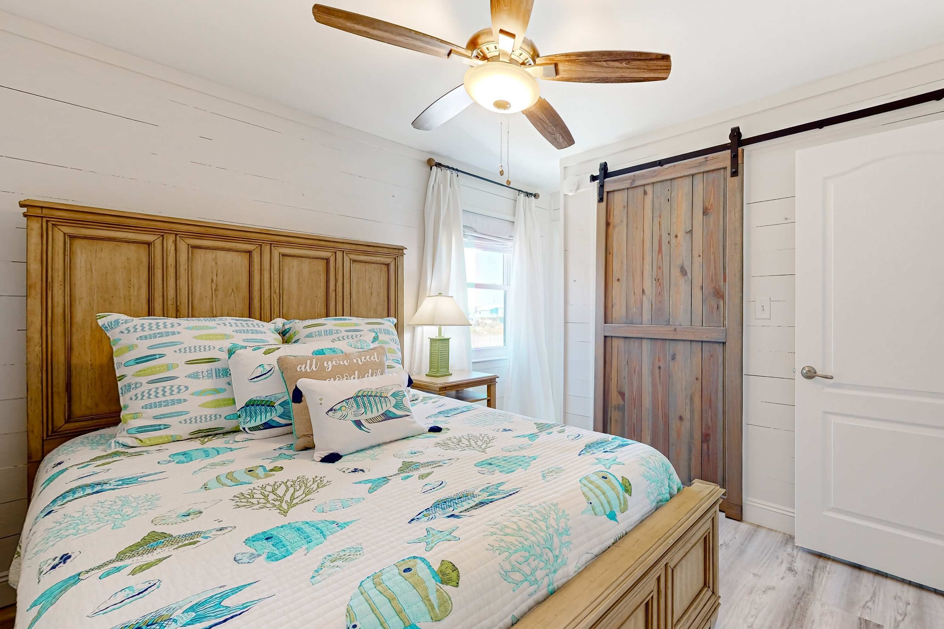 Een slaapkamer met een houten hoofdeinde, een bed met een marine-thema sprei, een plafondventilator en een schuifdeur in boerderijstijl.