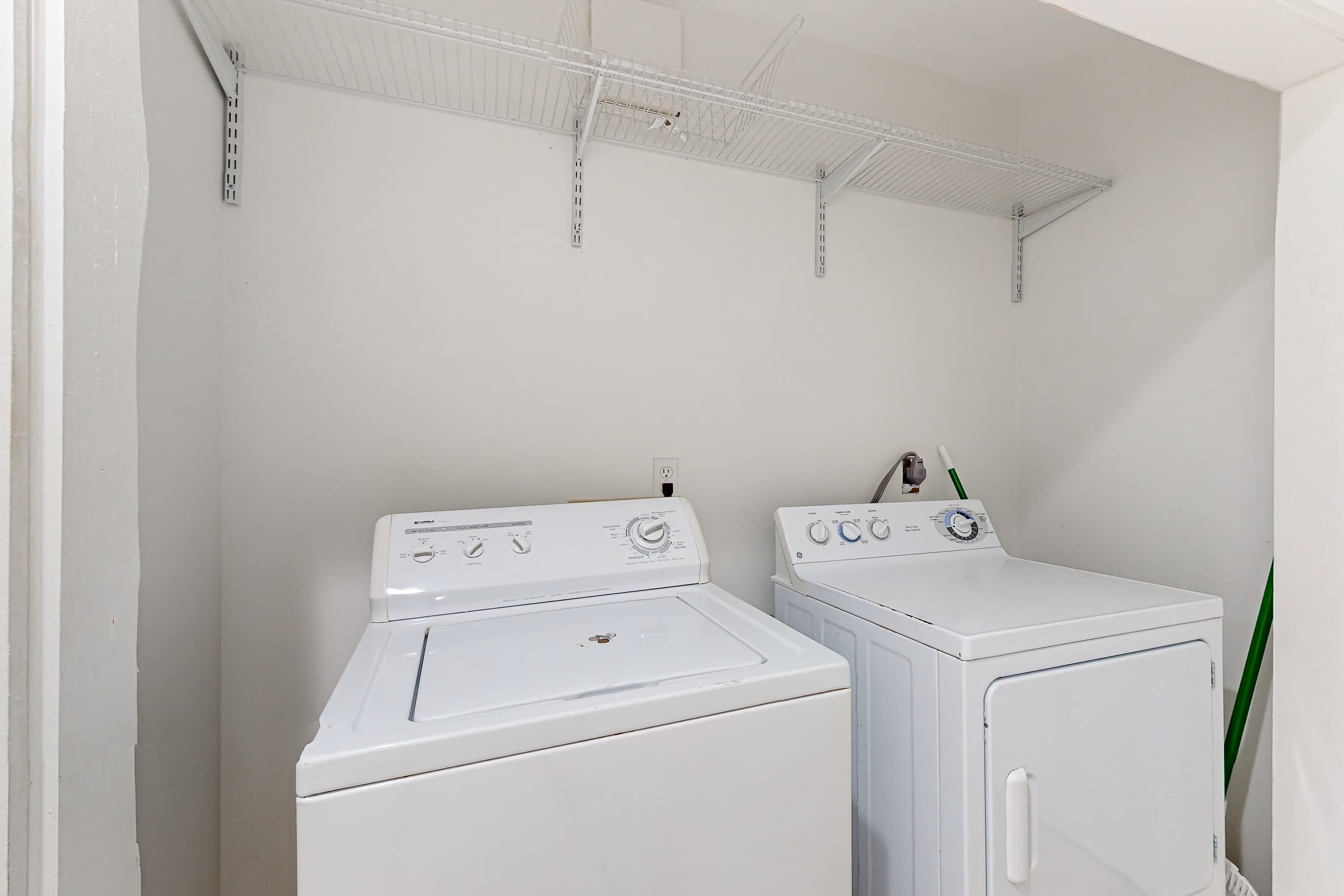 A laundry area with two white washing machines, a metal shelf above, and a green plant.