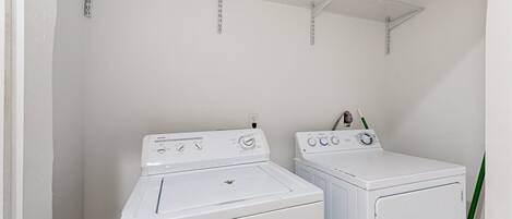 A laundry area with two white washing machines, a metal shelf above, and a green plant.