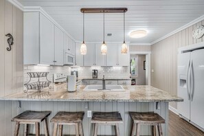 A kitchen with white cabinets, a marble countertop, hanging pendant lights, and a built-in island with stools. - Charming bay front home with WiFi, central AC, balcony, & deck (Orange Beach)