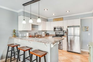 A kitchen with white cabinets, a stainless steel refrigerator, a built-in oven, and a granite countertop island with bar stools. - Convenient and Comfortable Suite in Beachfront Resort w/Shared Pool, Free WiFi (Orange Beach)