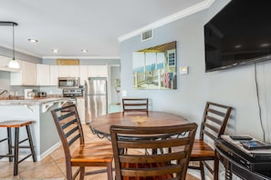 A modern kitchen with white cabinets, a dining table with wooden chairs, a TV mounted on the wall, and a painting above the table. - Convenient and Comfortable Suite in Beachfront Resort w/Shared Pool, Free WiFi (Orange Beach)