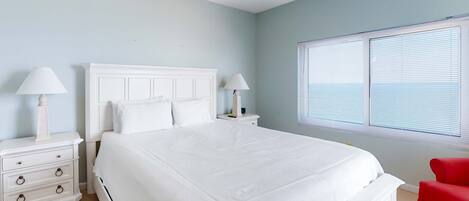 A bedroom with a large bed, white nightstands, two lamps, a red chair, and a window with blinds.