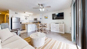 A modern kitchen with white cabinets, a dining area with a wooden table and chairs, a ceiling fan, and a flat-screen TV mounted on the wall. - Excellent resort suite in beachfront location with WiFi, pool & elevator (Orange Beach)