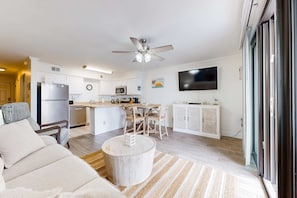 A modern kitchen with white cabinets, a dining area with a wooden table and chairs, a ceiling fan, and a flat-screen TV mounted on the wall. - Excellent resort suite in beachfront location with WiFi, pool & elevator (Orange Beach)