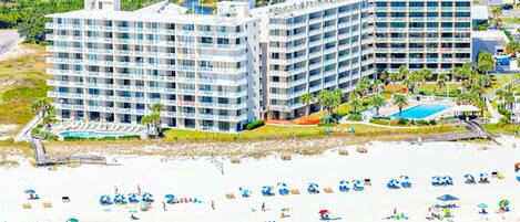 A beachfront hotel with multiple floors, a pool, and a beach with umbrellas.