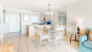 Amplia sala de estar con mesa de vidrio, sillas blancas, sofá, lámpara y cocina al fondo.