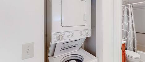 Un espacio pequeño para la lavandería con lavadora y secadora de carga frontal, un enchufe visible y una cortina de ducha en el baño contiguo.