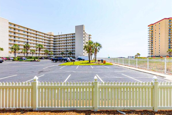 A multi-story hotel with a parking lot, palm trees, and a white picket fence. - Perfectly located condo in beachfront complex with pool, sauna & WiFi (Orange Beach)